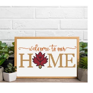 May include: A framed cross-stitch with the words "welcome to our HOME". The word HOME is in large, tan letters with a red maple leaf in the center. The text is stitched on white fabric, set in a wooden frame.