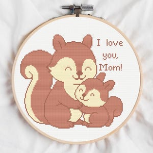 May include: Cross-stitch embroidery in a wooden hoop features two hugging squirrels in shades of brown and cream. The text "I love you, Mom!" is stitched above the squirrels. The background is white.