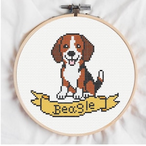 May include: A cross-stitch embroidery hoop featuring a smiling beagle dog. The dog is brown, black, and white, sitting above a yellow banner with the word "Beagle" stitched in black. The hoop is made of wood and set against a white fabric background.