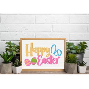 Könnte beinhalten: Ein gerahmtes Kreuzstichdesign mit den Worten "Happy Easter" in Gelb und Rosa, zusammen mit blauen Hasenohren und zwei verzierten Ostereiern. Das Design befindet sich vor einer weißen Backsteinmauer, mit Topfpflanzen im Vordergrund.