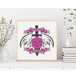 Può includere: Opera d'arte a punto croce incorniciata in una cornice dorata. Il design presenta un dado D20 rosa, ali, una spada e banner con la scritta "Dungeon Mistress". Lo sfondo è bianco. L'opera è esposta su una superficie bianca.