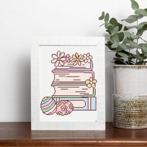 May include: A framed cross-stitch design featuring a stack of books adorned with flowers. Two decorative Easter eggs are in front of the books. The color palette includes pastel pink, yellow, green, and blue.