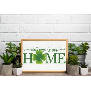 Op de afbeelding: Een ingelijst kruissteekpatroon met de tekst "welcome to our HOME" in het groen, waarbij een klavertjevier de letter "O" vervangt. Het ontwerp is ingelijst in een lichthouten lijst en omringd door verschillende kleine vetplantjes in potjes.