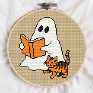 May include: A circular wooden embroidery hoop containing a cross-stitch pattern on tan fabric. The design features a white ghost with black eyes holding an open orange book, accompanied by an orange and black striped tabby cat standing below.