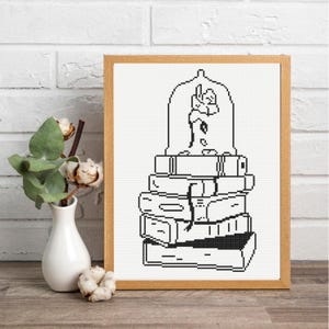 May include: A framed cross-stitch pattern featuring a black and white pixel art design of a single rose enclosed within a glass dome, resting atop a vertical stack of five books, presented in a light-colored wooden frame with a clean aesthetic.