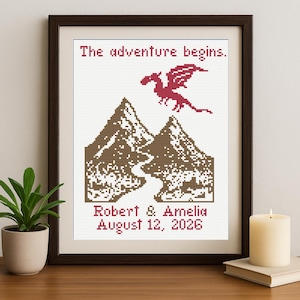 May include: Cross-stitch artwork in a dark wood frame. The design features two brown mountains, a red dragon, and the text "The adventure begins." Below, it reads "Robert & Amelia August 12, 2026."