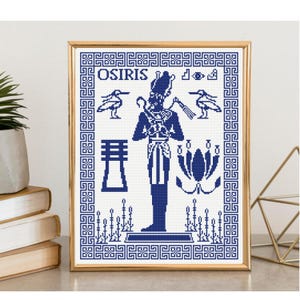 Puede incluir: Un patrón de punto de cruz enmarcado en un marco dorado con un diseño azul y blanco de la deidad egipcia Osiris. La obra incluye el texto OSIRIS, dos pájaros, un pilar djed, una flor de loto y un patrón de borde geométrico.