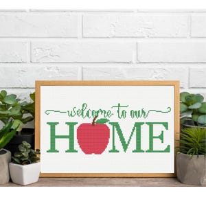 Może przedstawiać: Oprawiona praca haftu krzyżykowego ze słowami "welcome to our HOME" w kolorze zielonym i czerwonym jabłkiem zastępującym "O". Grafika jest w jasnobrązowej drewnianej ramie i jest wyświetlana na tle białej ceglanej ściany.