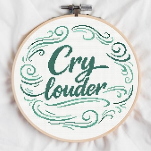 May include: A round, wooden embroidery hoop holds a cross-stitch design. The text "Cry louder" is stitched in a dark teal green, surrounded by decorative swirls in a lighter shade of green. The background is white.
