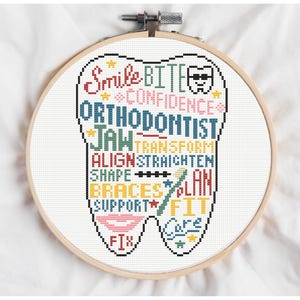 Op de afbeelding: Een kruissteekborduurwerk in een houten hoepel. Het ontwerp is een tandvorm gevuld met woorden als "Smile", "Orthodontist" en "Braces". Het kleurenpalet omvat rood, blauw, geel en groen. Het algemene thema is tandheelkundige gezondheid.