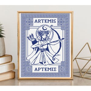 May include: A framed cross-stitch pattern featuring a blue and white design of the goddess Artemis holding a bow and arrow. The artwork includes a crescent moon, a floral crown, and the text ARTEMIS in English and Greek characters, all enclosed within a geometric Greek key border.