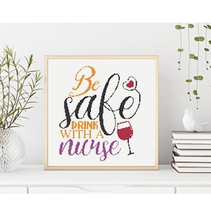 Op de afbeelding: Een ingelijst kruissteekkunstwerk met de woorden "Be safe drink with a nurse" in oranje, zwart en paars. Een rood wijnglas is te zien in het ontwerp. Het kunstwerk wordt tentoongesteld op een wit oppervlak met een vaas groen en een stapel boeken.
