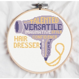 Puede incluir: Un bastidor de bordado de punto de cruz con un diseño de secador de pelo morado y amarillo. El diseño incluye las palabras "TALENTED", "VERSATILE", "IMAGINATIVE", "STYLE", "CONFIDENCE", "HAIR DRESSER" y "ARTIST". El bordado está sobre un fondo blanco.