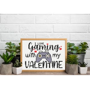 Może przedstawiać: Oprawiony haft krzyżykowy z napisem "I Love Gaming with my Valentine" i szarym kontrolerem do gier. Grafika jest w drewnianej ramie i jest otoczona roślinami doniczkowymi.