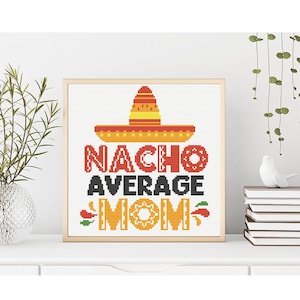 May include: Cross-stitch art in a wooden frame. The design features a sombrero above the words "NACHO AVERAGE MOM" in red, black, and yellow. The artwork is displayed on a white surface with decorative plants and books.