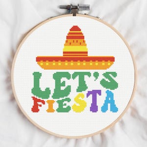 May include: A cross-stitch embroidery hoop featuring a colorful sombrero and the words "LET'S FIESTA" in various colors. The design is stitched on white fabric and framed in a wooden hoop.