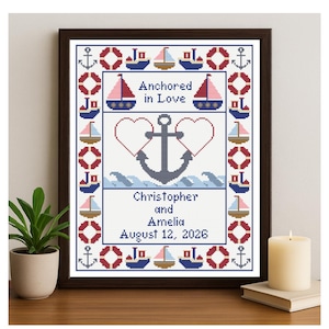 May include: Cross-stitch artwork in a dark wood frame. The design features an anchor, hearts, sailboats, and the text "Anchored in Love," along with names and a date. The color palette includes red, blue, and white.