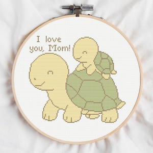 Puede incluir: Bordado de punto de cruz en un aro de madera con dos tortugas, una más grande que lleva a otra más pequeña. El texto "I love you, Mom!" está bordado encima de las tortugas. Las tortugas son de color amarillo claro y verde.
