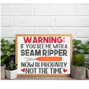 May include: A framed cross-stitch with the text "WARNING: IF YOU SEE ME WITH A SEAM RIPPER NOW IS PROBABLY NOT THE TIME." The text is in black and red, with an orange seam ripper graphic. The frame is light brown.