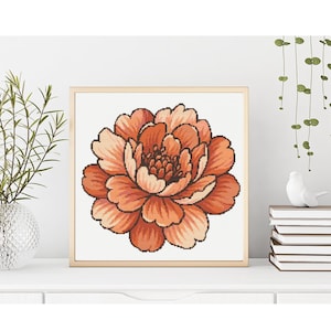 May include: A framed cross-stitch of a large peony flower in shades of orange and peach. The flower is centered on a white background, with a gold-colored frame. The artwork is displayed on a white surface.