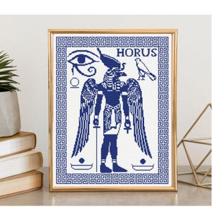 Puede incluir: Un patrón de punto de cruz enmarcado que presenta una representación en azul oscuro de la deidad egipcia Horus con alas, el Ojo de Horus y un icono de halcón. El diseño está encerrado en un borde geométrico griego con el texto HORUS arriba.