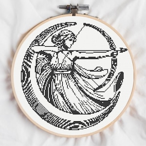 May include: A circular wooden embroidery hoop containing a black and white cross-stitch pattern of a figure with long hair holding a bow and arrow, positioned within a large crescent moon shape, featuring detailed linework and pixelated textures.