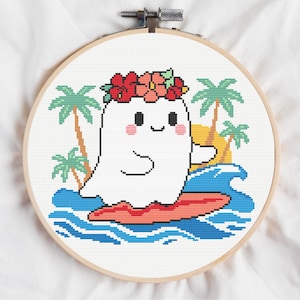 May include: Cross-stitch embroidery in a wooden hoop. The design features a smiling white ghost wearing a flower crown, surfing on a red surfboard. Palm trees, waves, and a yellow sun complete the beach scene.