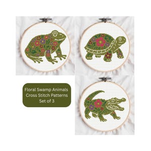 May include: Three cross-stitch patterns featuring swamp animals: a frog, a turtle, and an alligator. Each animal is green and adorned with floral patterns in red, purple, and yellow. The patterns are set in wooden embroidery hoops.