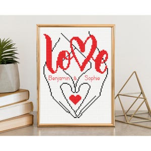 Peut inclure: Une œuvre d'art au point de croix encadrée avec le mot "Love" en script rouge, représentant un cœur et les noms "Benjamin & Sophie". L'œuvre est dans un cadre doré, avec un fond blanc et des points noirs.