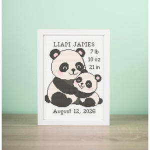 May include: A framed cross-stitch featuring two pandas, one embracing the other. The text reads "LIAM JAMES" above the baby's birth details: 7 lb, 10 oz, and 21 in. The date "August 12, 2026" is below the pandas. The frame is white.