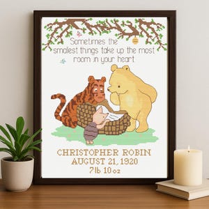 May include: Framed cross-stitch art depicting Winnie the Pooh characters. The text reads "Sometimes the smallest things take up the most room in your heart." Below, the name "Christopher Robin" and birth details are displayed. The frame is dark wood.