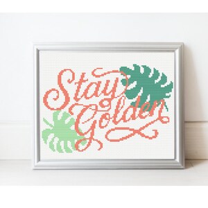 May include: A framed cross-stitch artwork with the words "Stay Golden" in coral-colored thread. The design includes green monstera leaves. The artwork is in a silver frame and set against a white background.