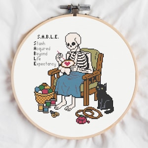 May include: Cross-stitch embroidery in a wooden hoop. The design features a skeleton seated in a chair, cross-stitching a heart. A black cat sits nearby. Text reads "S.A.B.L.E. Stash Acquired Beyond Life Expectancy."