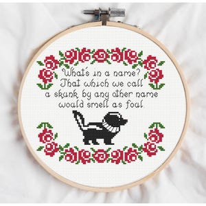 May include: Cross-stitch embroidery in a wooden hoop. The design features a black and white skunk with a floral border of red roses and green leaves. Text reads, "What's in a name? That which we call a skunk by any other name would smell as foul."