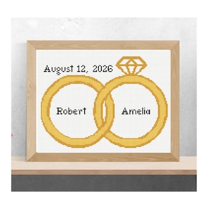 Puede incluir: Obra de arte de punto de cruz en un marco de madera. El diseño presenta dos anillos de boda dorados entrelazados con los nombres "Robert" y "Amelia" en el interior. Encima de los anillos está la fecha "12 de agosto de 2026" y un diseño en forma de diamante.