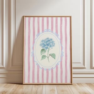 May include: A framed art print featuring a watercolor illustration of a blue hydrangea flower. The artwork is set against a background of pink and white vertical stripes and framed in a gold-colored frame. The floral art is in an oval frame.