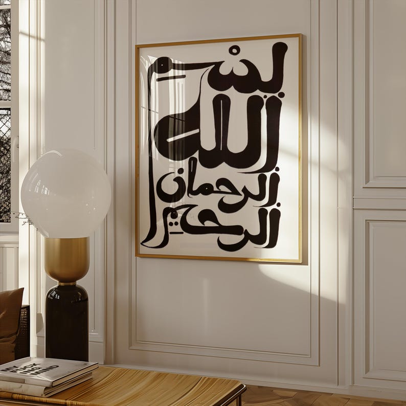 A framed art print featuring black Arabic calligraphy on a white background. The artwork is encased in a gold frame and hangs on a white wall. The print's design is a bold, modern interpretation of traditional calligraphy.