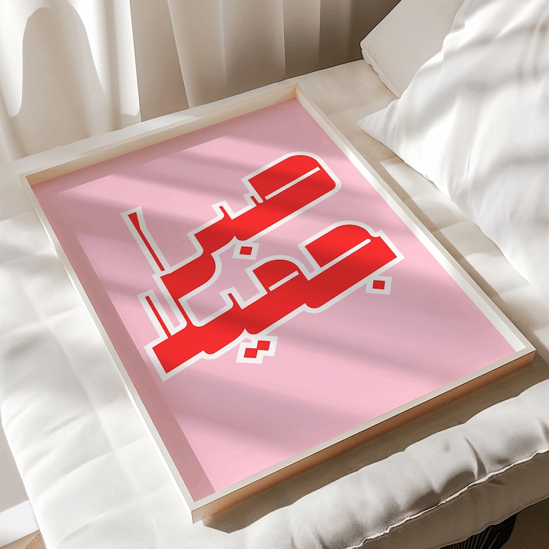 May include: A framed art print featuring Arabic calligraphy in red against a pink background. The artwork is encased in a white frame, creating a modern and minimalist aesthetic. The print is displayed on a white surface.