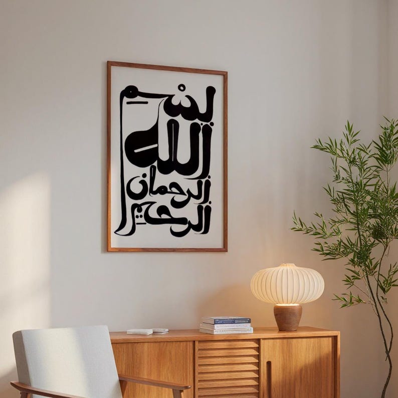 A framed art print featuring black Arabic calligraphy on a white background. The artwork is in a wooden frame and hangs on a white wall. The room also includes a wooden cabinet, a lamp, and a plant.