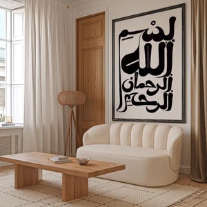 A living room scene featuring a large framed artwork with Arabic calligraphy in black against a cream background. A cream-colored sofa, wooden coffee table, and a woven lamp are also visible. The room has a neutral color palette.