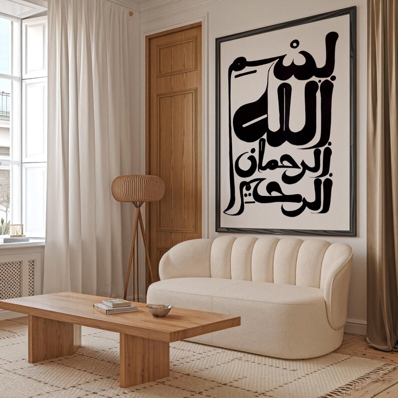A living room scene featuring a large framed artwork with Arabic calligraphy in black against a cream background. A cream-colored sofa, wooden coffee table, and a woven lamp are also visible. The room has a neutral color palette.