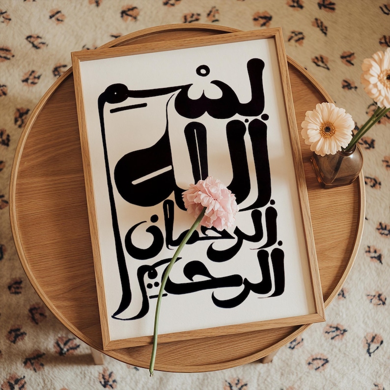 A framed art print featuring black Arabic calligraphy on a white background. The wooden frame surrounds the artwork, which is placed on a round wooden table. A pink flower rests on the print, and a small vase with flowers is to the right.