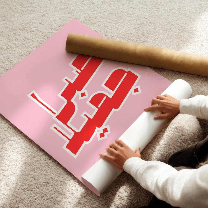 May include: A pink poster with red Arabic calligraphy is unrolled on a light-colored carpet. A rolled-up poster is next to it. The poster's design features bold, stylized lettering.