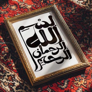 A framed print featuring Arabic calligraphy in black against a white background. The gold frame rests on a red patterned rug. The calligraphy is a stylized script.