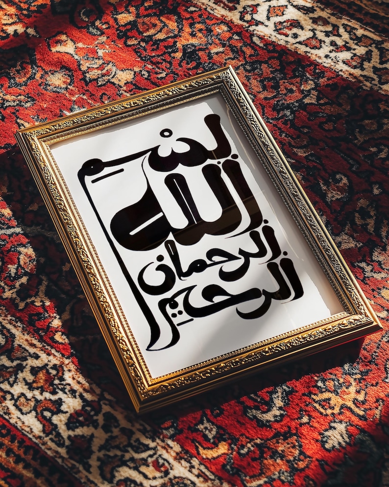 A framed print featuring Arabic calligraphy in black against a white background. The gold frame rests on a red patterned rug. The calligraphy is a stylized script.