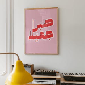 May include: A framed pink art print with red Arabic calligraphy. The artwork is in a light wood frame and hangs on a white wall. A yellow lamp and audio equipment are in the foreground.
