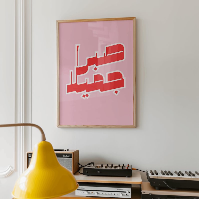 May include: A framed pink art print with red Arabic calligraphy. The artwork is in a light wood frame and hangs on a white wall. A yellow lamp and audio equipment are in the foreground.