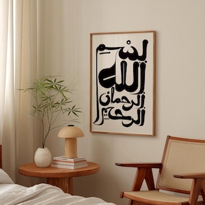 A framed art print featuring black Arabic calligraphy on a cream background. The artwork is in a wooden frame and hangs on a neutral-colored wall. The room also includes a wooden side table with a lamp and a plant.