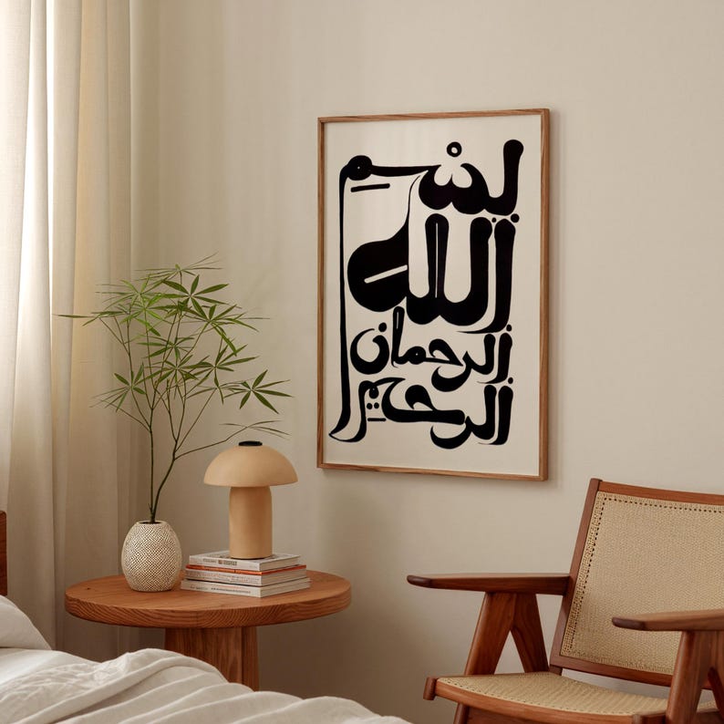 A framed art print featuring black Arabic calligraphy on a cream background. The artwork is in a wooden frame and hangs on a neutral-colored wall. The room also includes a wooden side table with a lamp and a plant.