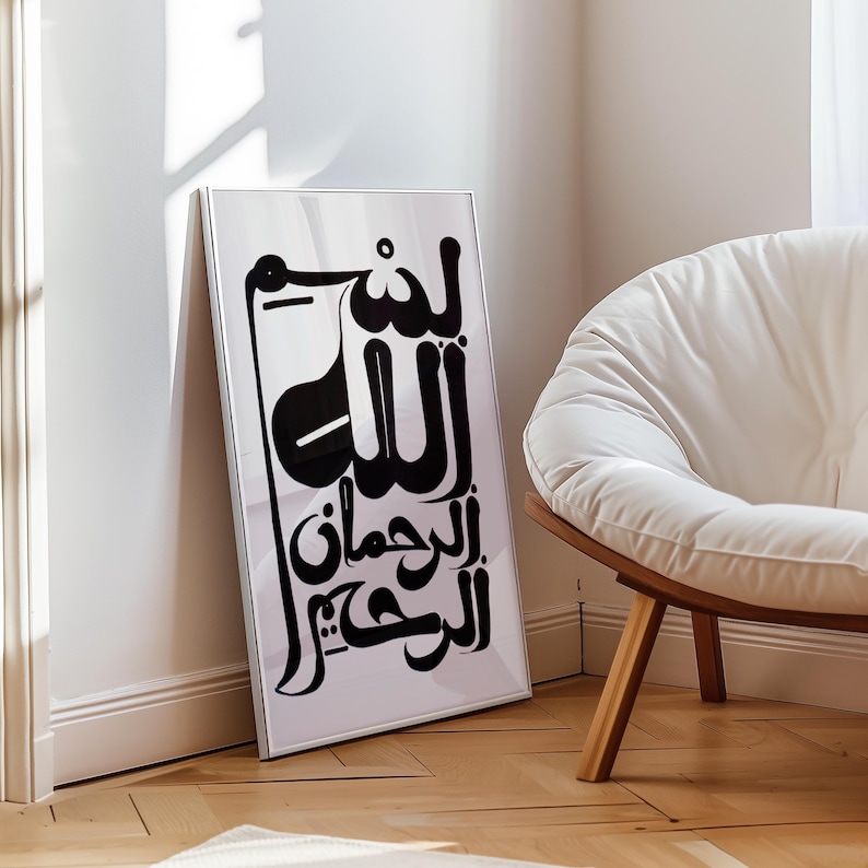 A framed black and white art print featuring Arabic calligraphy. The artwork leans against a white wall, with a modern white chair and wooden floor in the background. The calligraphy is in a bold, stylized font.